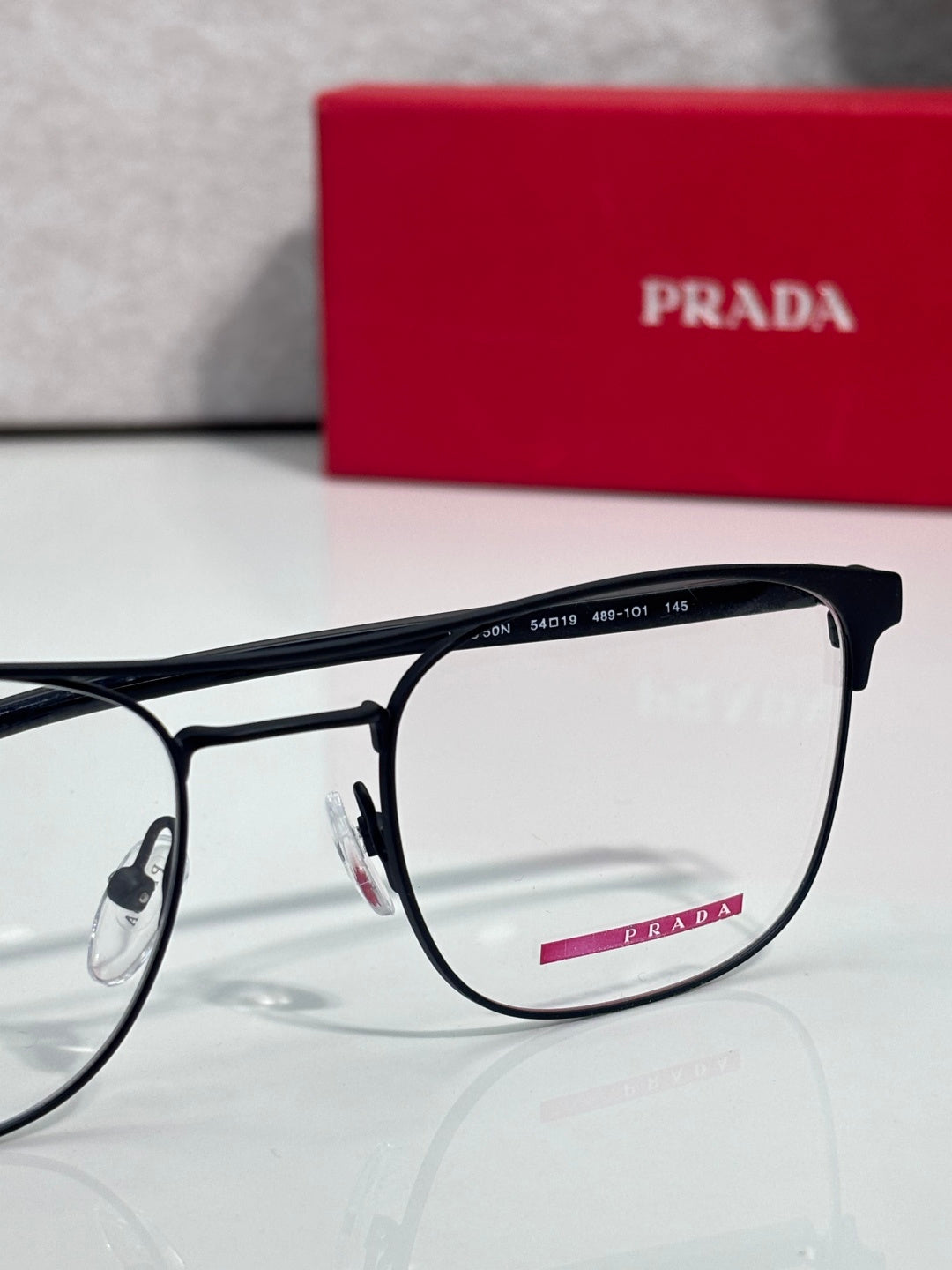 Black full-rim eyeglasses with clear lenses and red logo accents, in front of a Prada case.