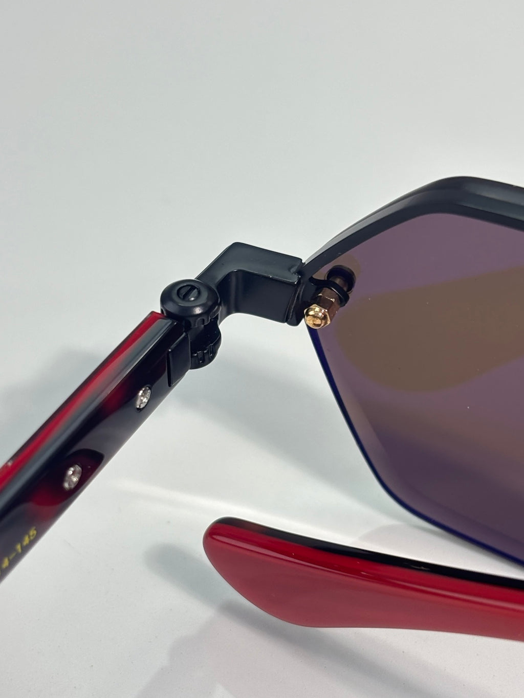 Close-up of hexagon sunglasses showing red temple arm and black hinge to tinted lens
