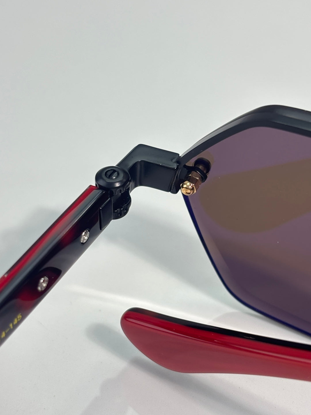 Close-up of hexagon sunglasses showing red temple arm and black hinge to tinted lens