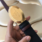 Prada belt close-up: gold oval PRADA MILANO buckle on dark strap.