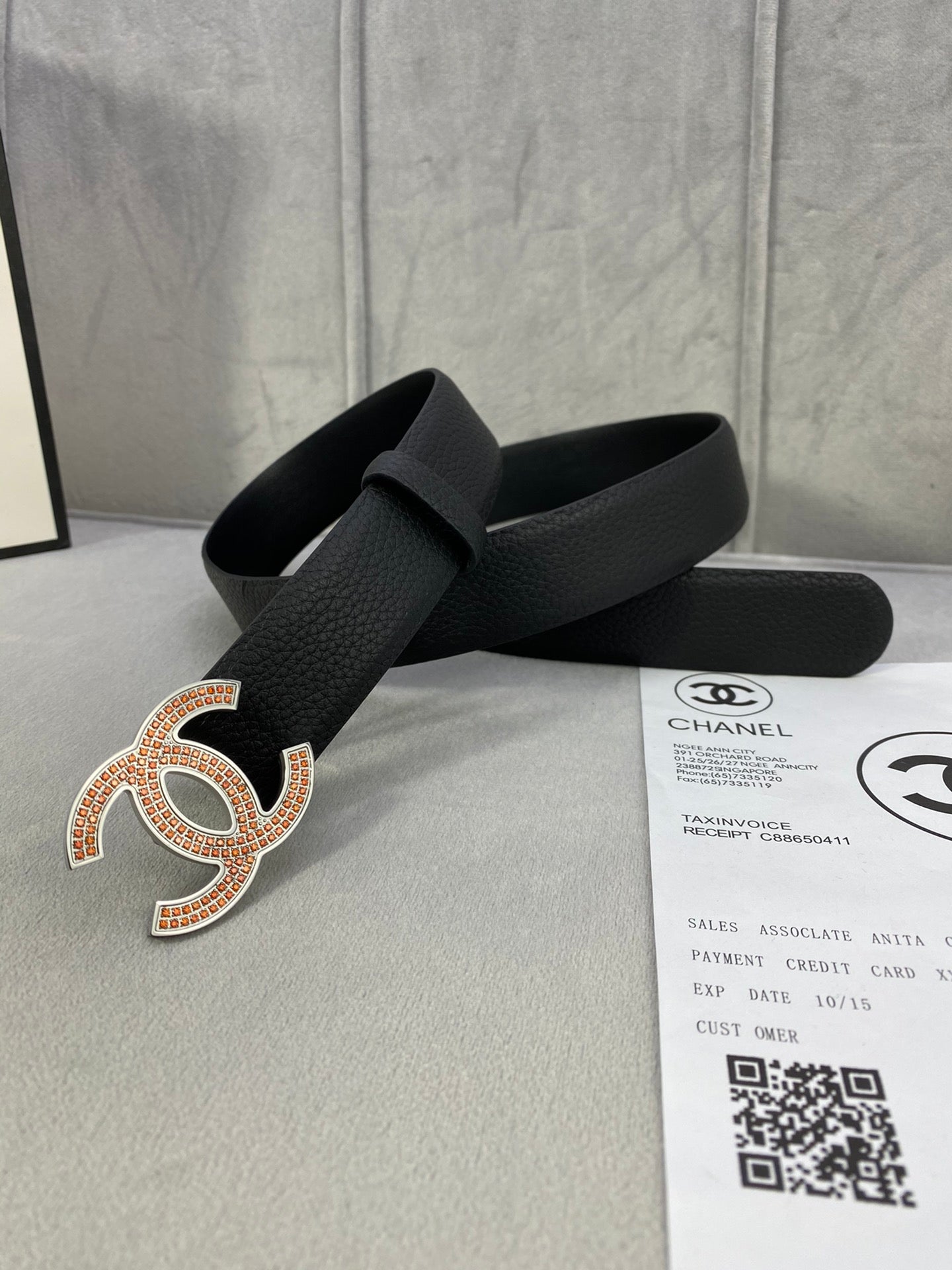 Black leather belt with gold and orange rhinestone CC logo buckle, strap shown over papers.