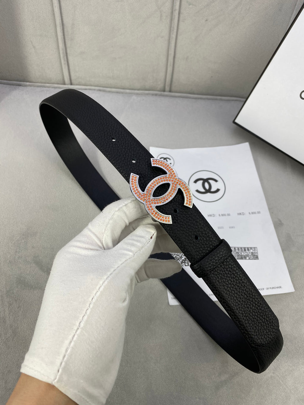 Black leather belt with orange and gold rhinestone CC logo buckle, laid over paper.