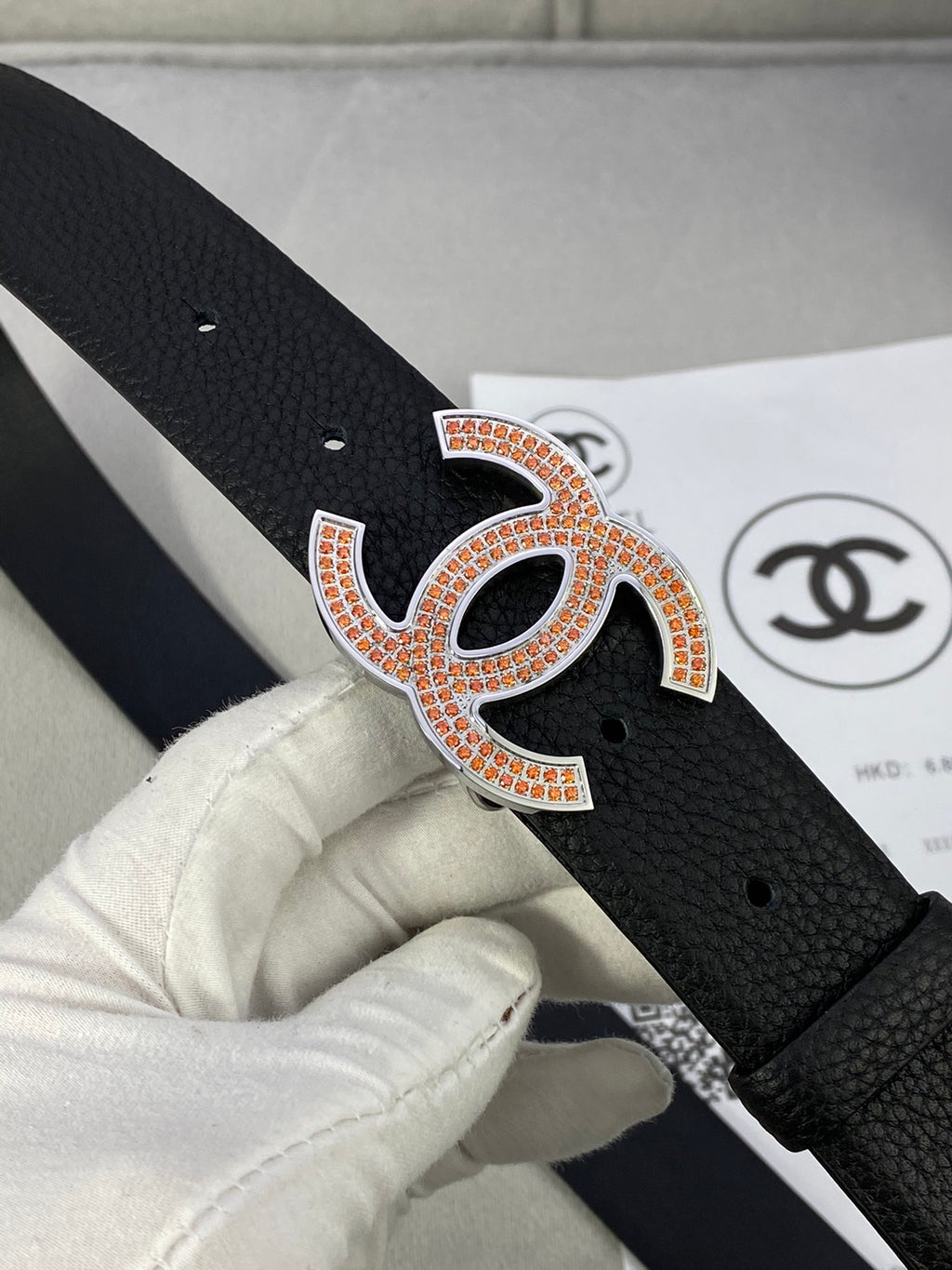 Close-up of black leather belt with orange-and-gold rhinestone CC logo buckle.