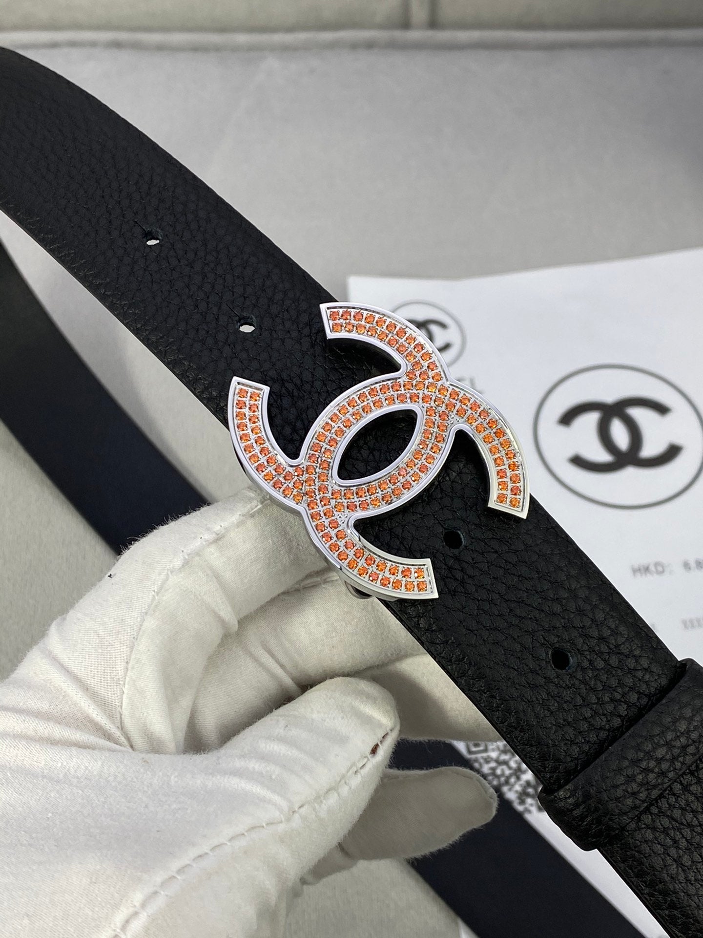 Close-up of black leather belt with orange-and-gold rhinestone CC logo buckle.