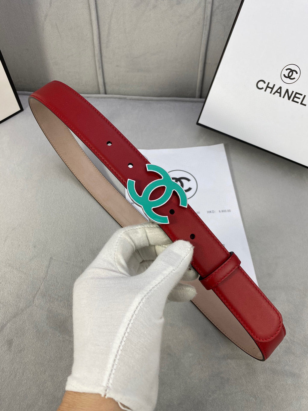 Red leather belt with teal CC-logo buckle, held in gloved hand