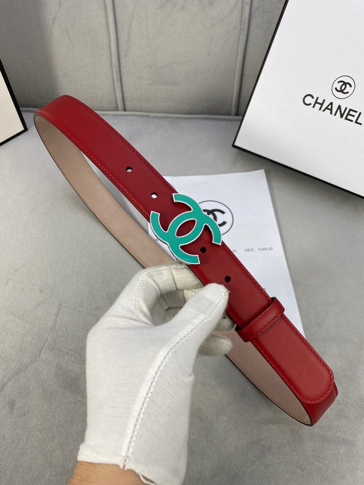Red leather belt with teal CC-logo buckle, held in gloved hand