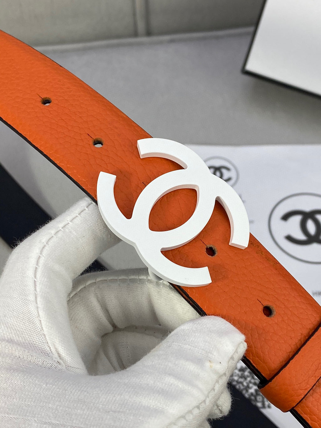 Chanel belt close-up with orange leather strap, perforation holes, and white CC buckle