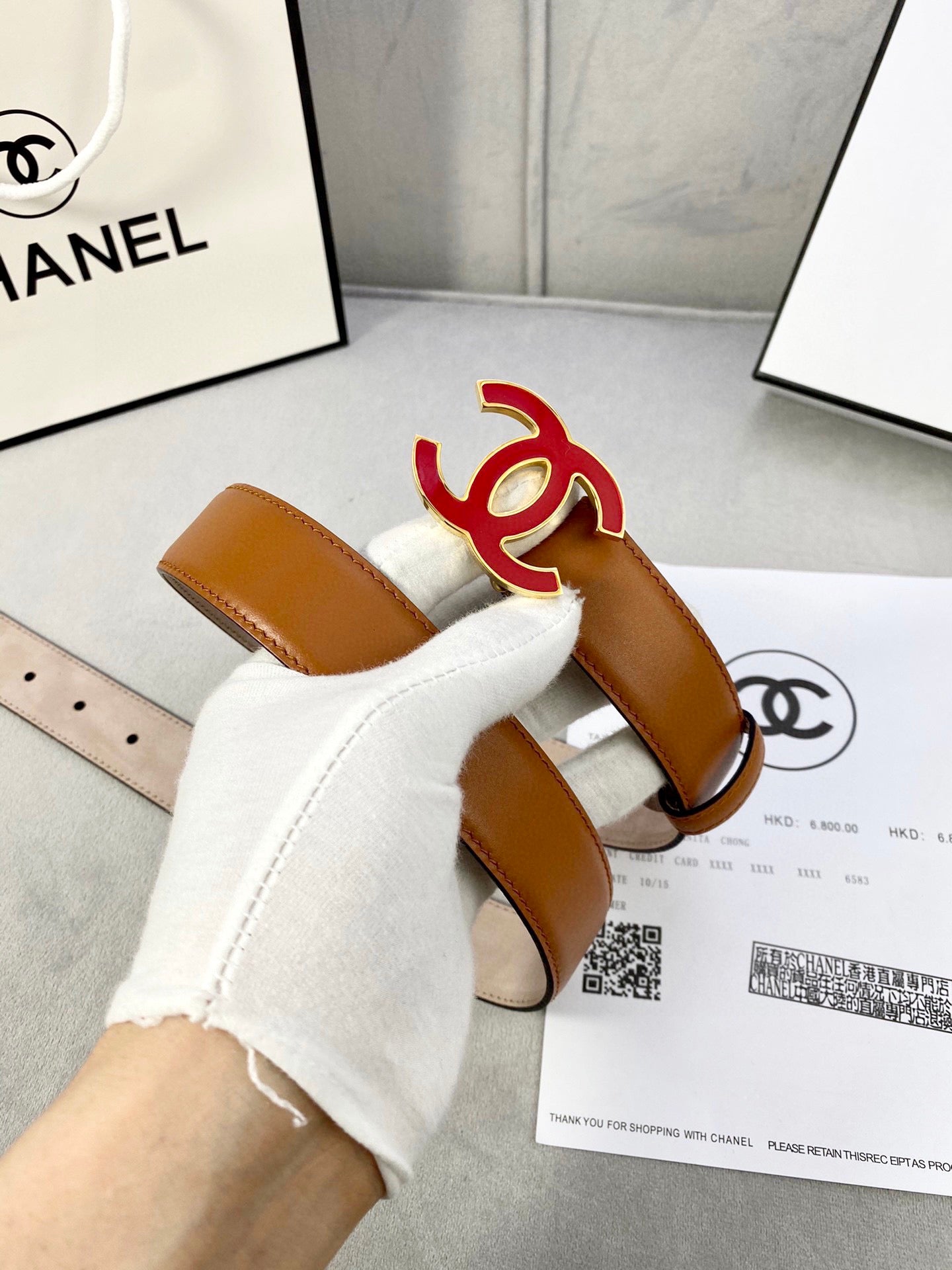 Brown Chanel belt held in gloved hand, featuring red CC emblem buckle