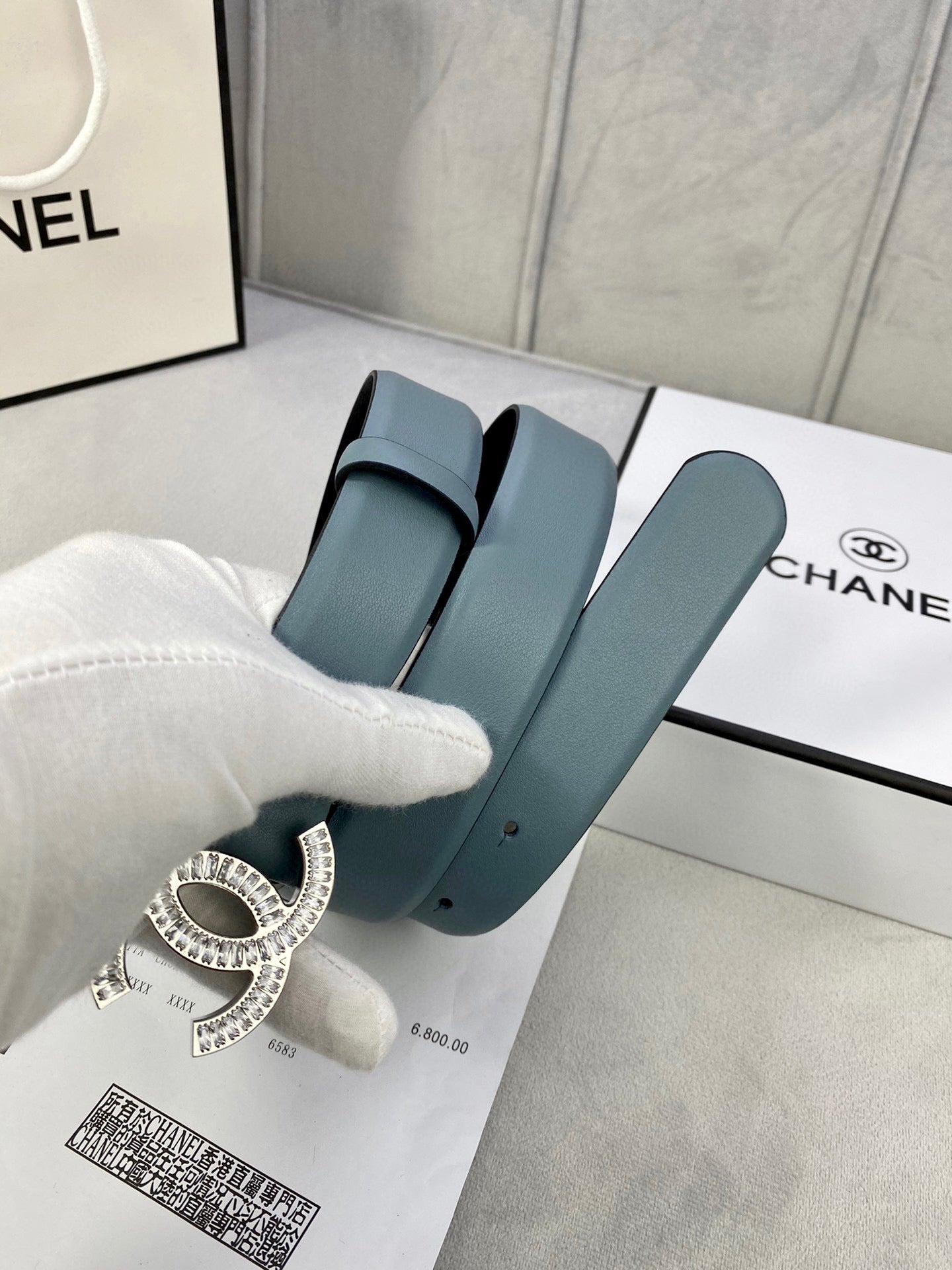 Light blue Chanel belt with silver-toned CC buckle over a shopping bag and box.