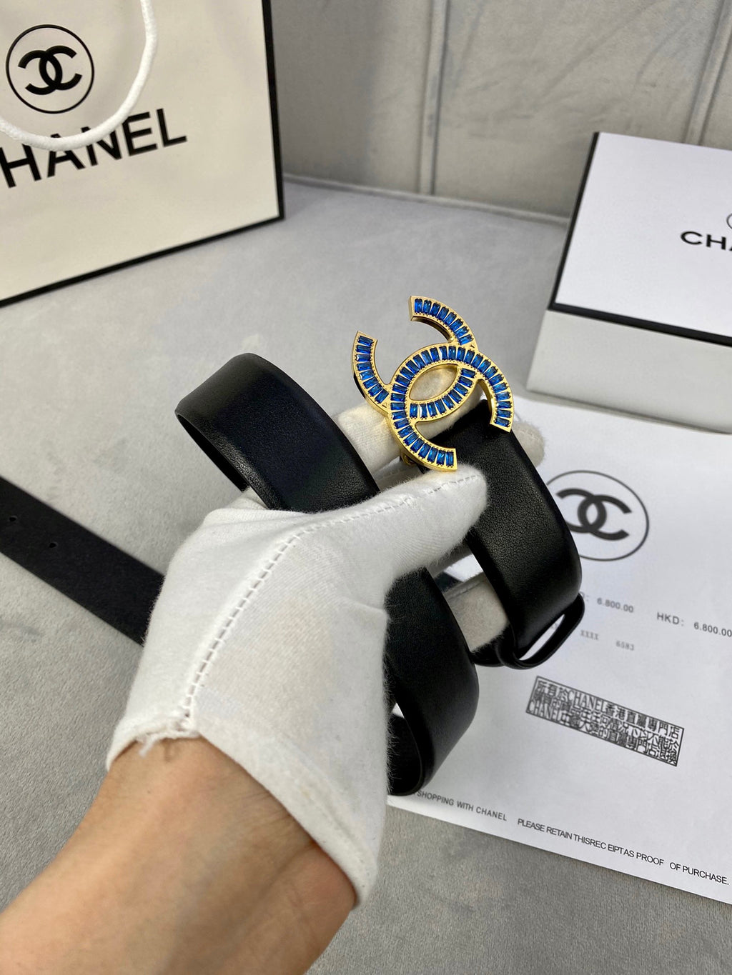 Chanel black leather belt with blue and gold CC logo buckle, held by glove