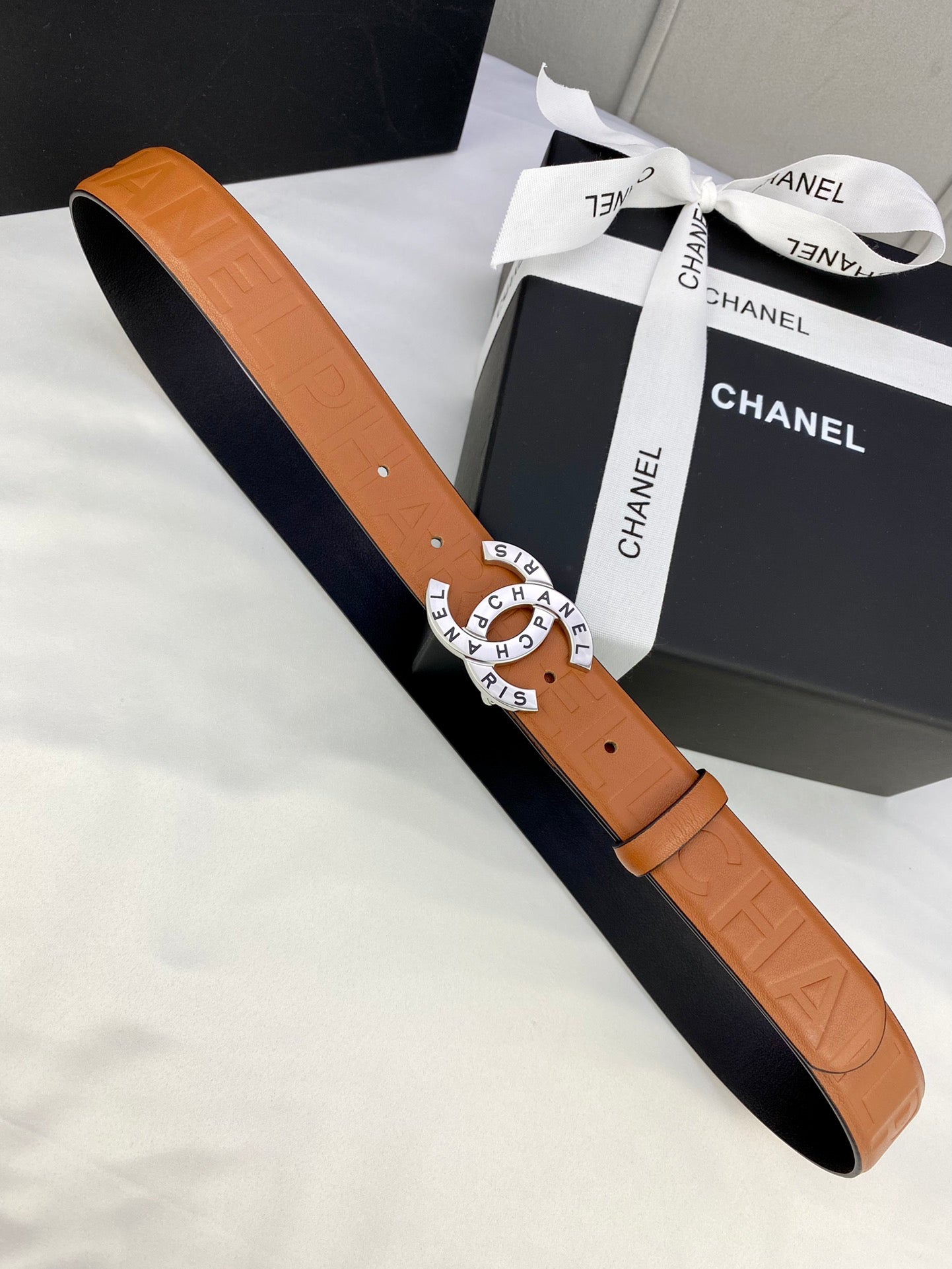 Tan-orange leather belt with embossed pattern, circular logo buckle on strap end.