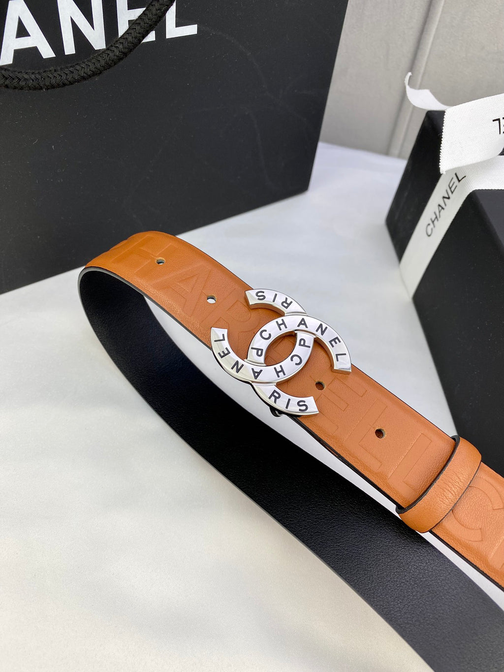 Tan-orange leather belt with circular logo buckle and visible strap edges.