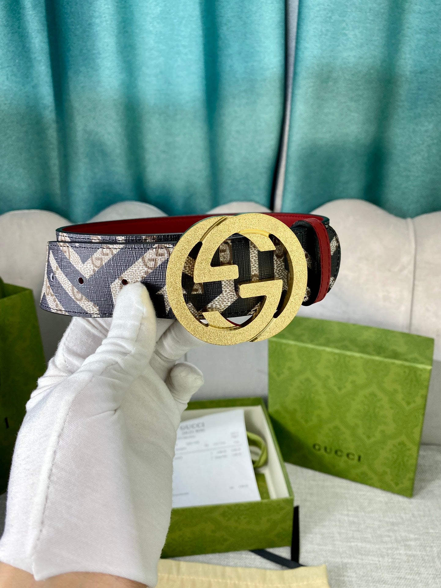 Logo-print belt with gold round buckle and red leather strap edge, held in white glove.