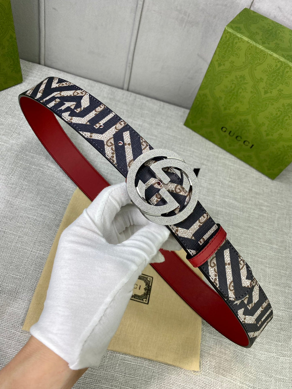 Logo-print belt with silver round G buckle and red leather underside, held by glove.