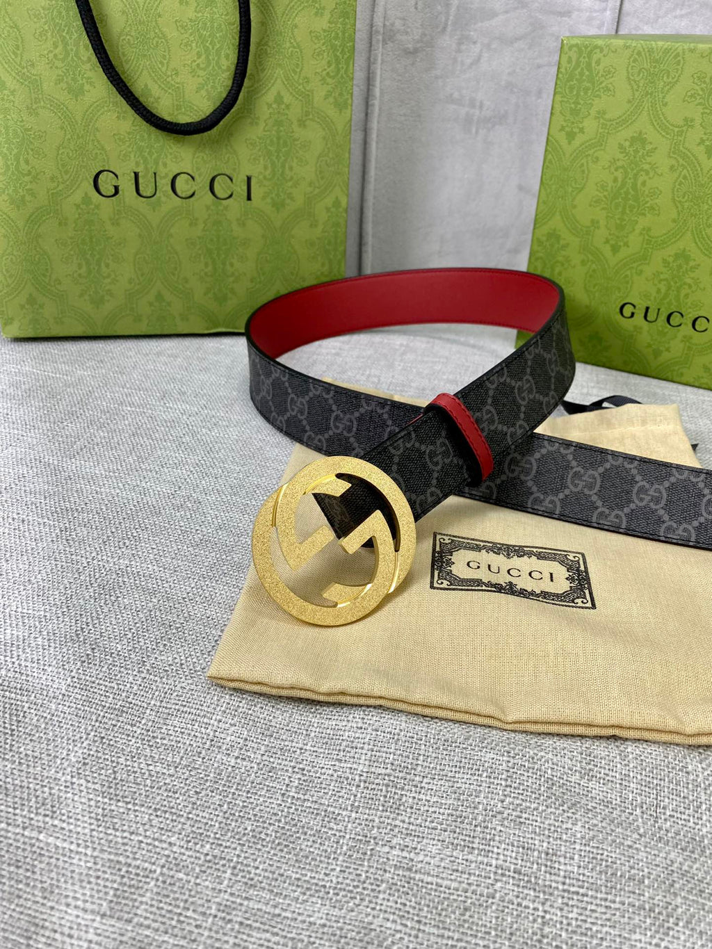 Designer belt on fabric surface: dark GG-print strap, red leather trim, and gold-tone buckle.