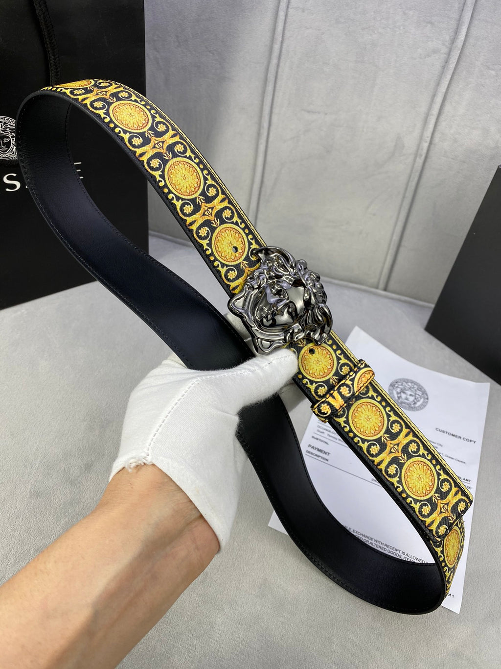 Designer belt held in hand: black strap, gold medallion trim, and silver lion buckle