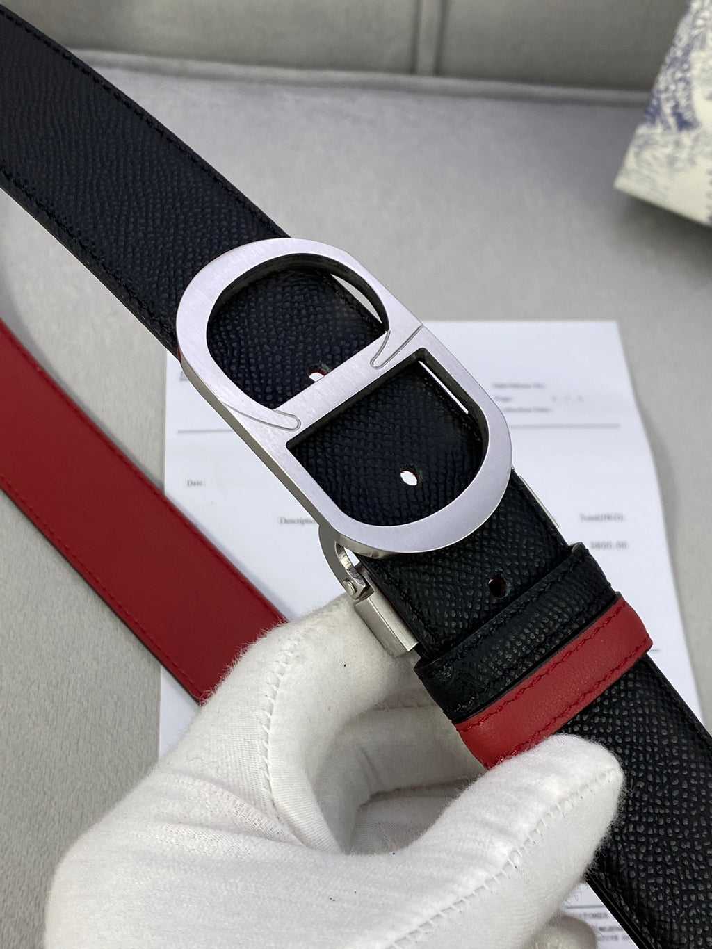 Dior belt buckle close-up: silver-tone oval frame on black strap with red edge.