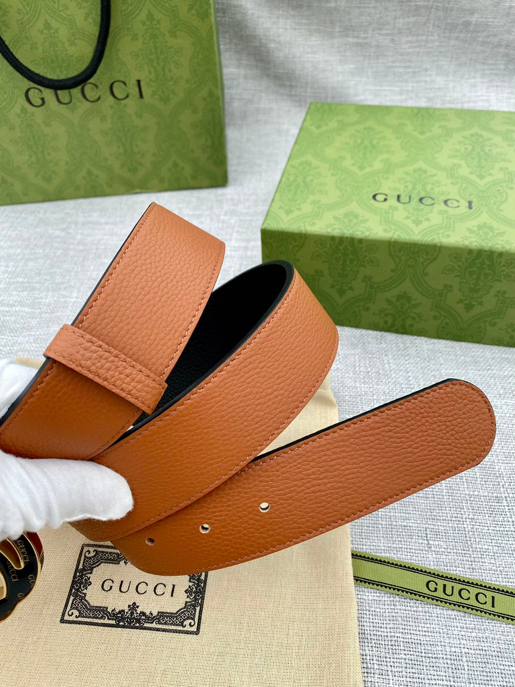 Tan leather belt with stitched edge and round metal logo buckle; strap with holes.