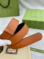 Tan leather belt with stitched edge and round metal logo buckle; strap with holes.