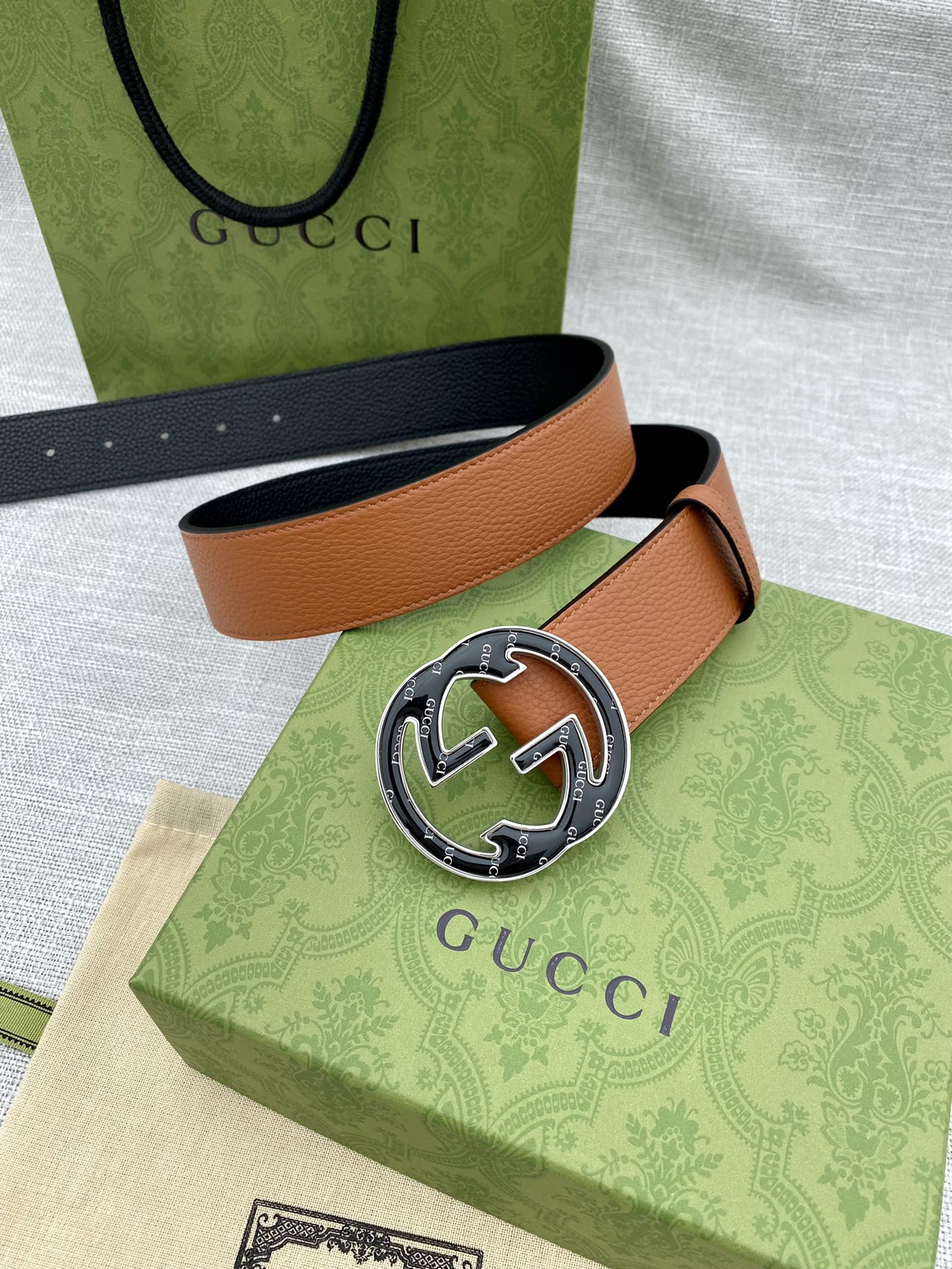 Tan leather belt with round logo buckle, laid across green and beige bags.
