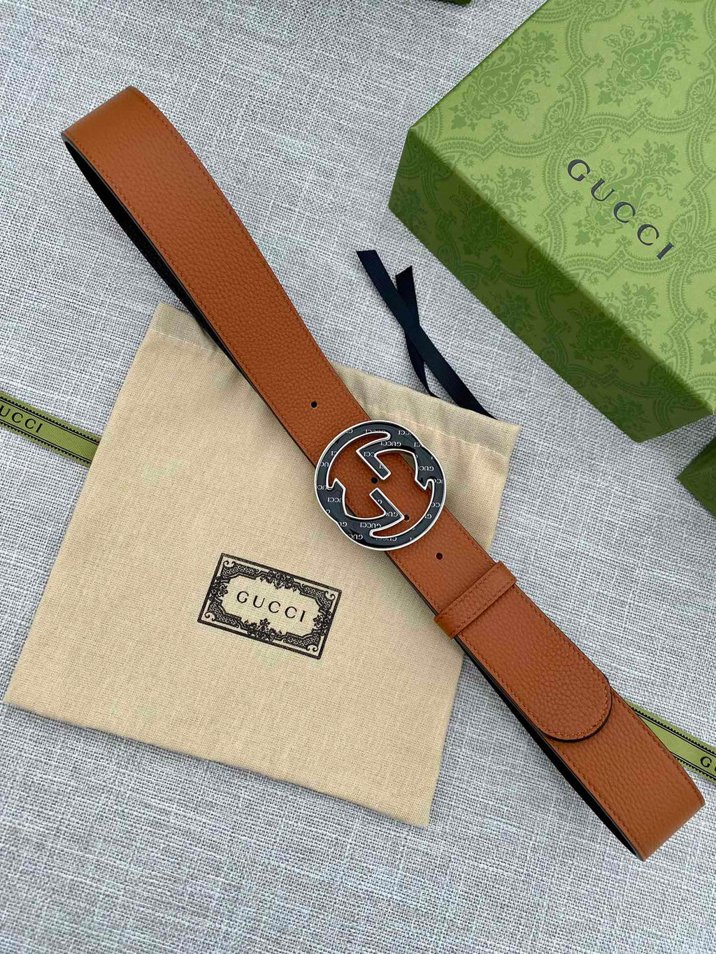 Tan leather belt with round logo buckle, laid flat on a beige cloth pouch.