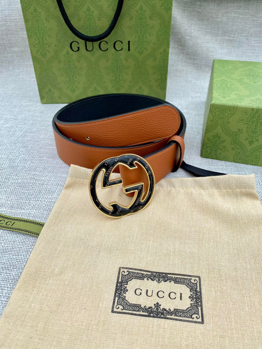 Tan leather belt with round logo buckle resting on beige pouch and packaging.