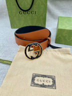 Tan leather belt with round logo buckle resting on beige pouch and packaging.