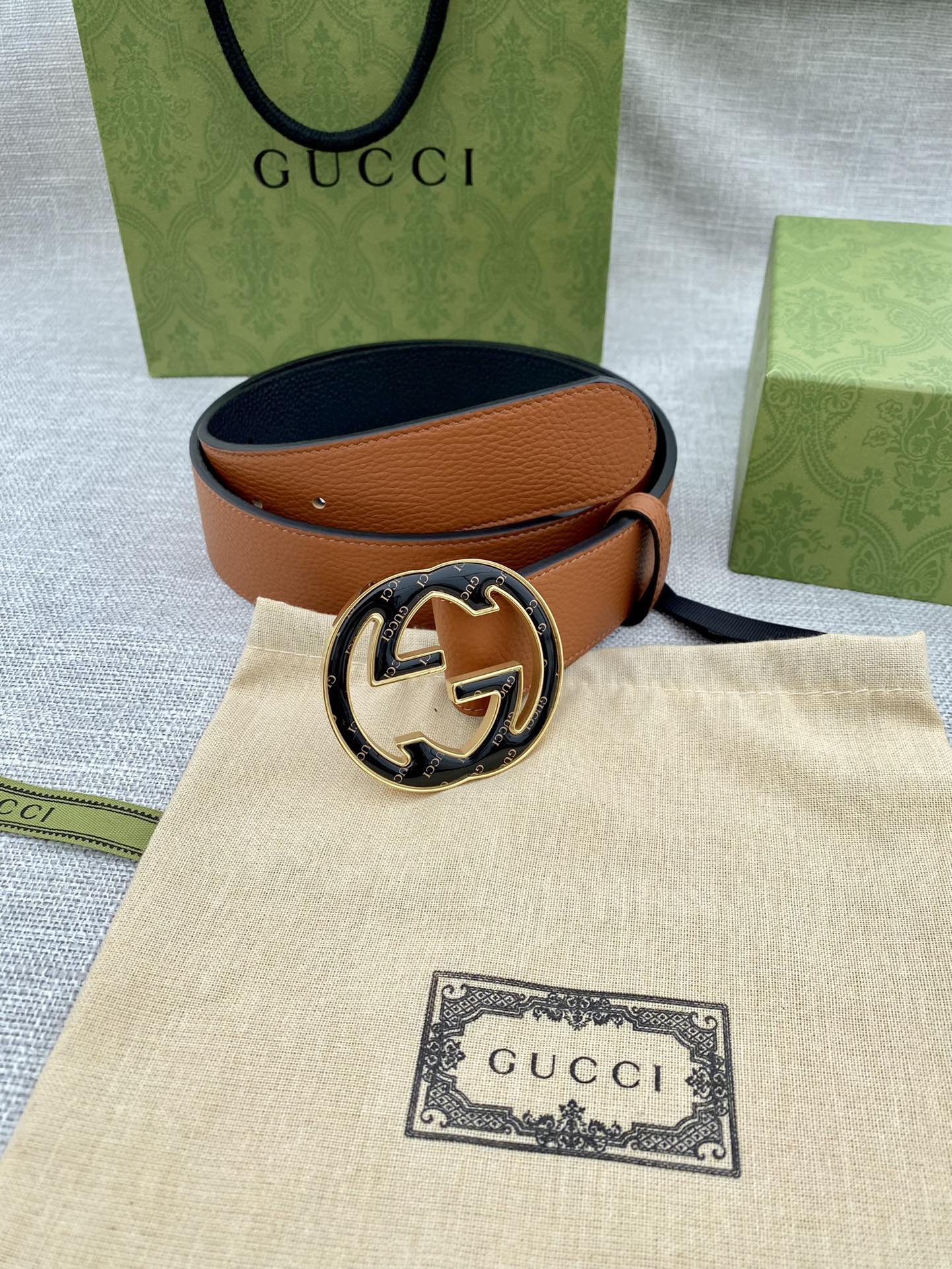 Tan leather belt with round logo buckle resting on beige pouch and packaging.