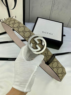 GG canvas belt with gold-tone double G buckle; held by gloved hand, strap underside visible.