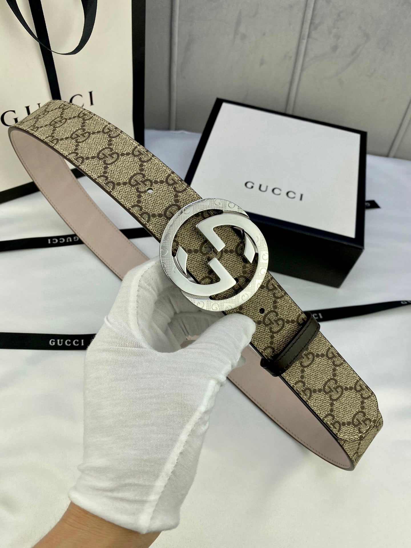 GG canvas belt with gold-tone double G buckle; held by gloved hand, strap underside visible.