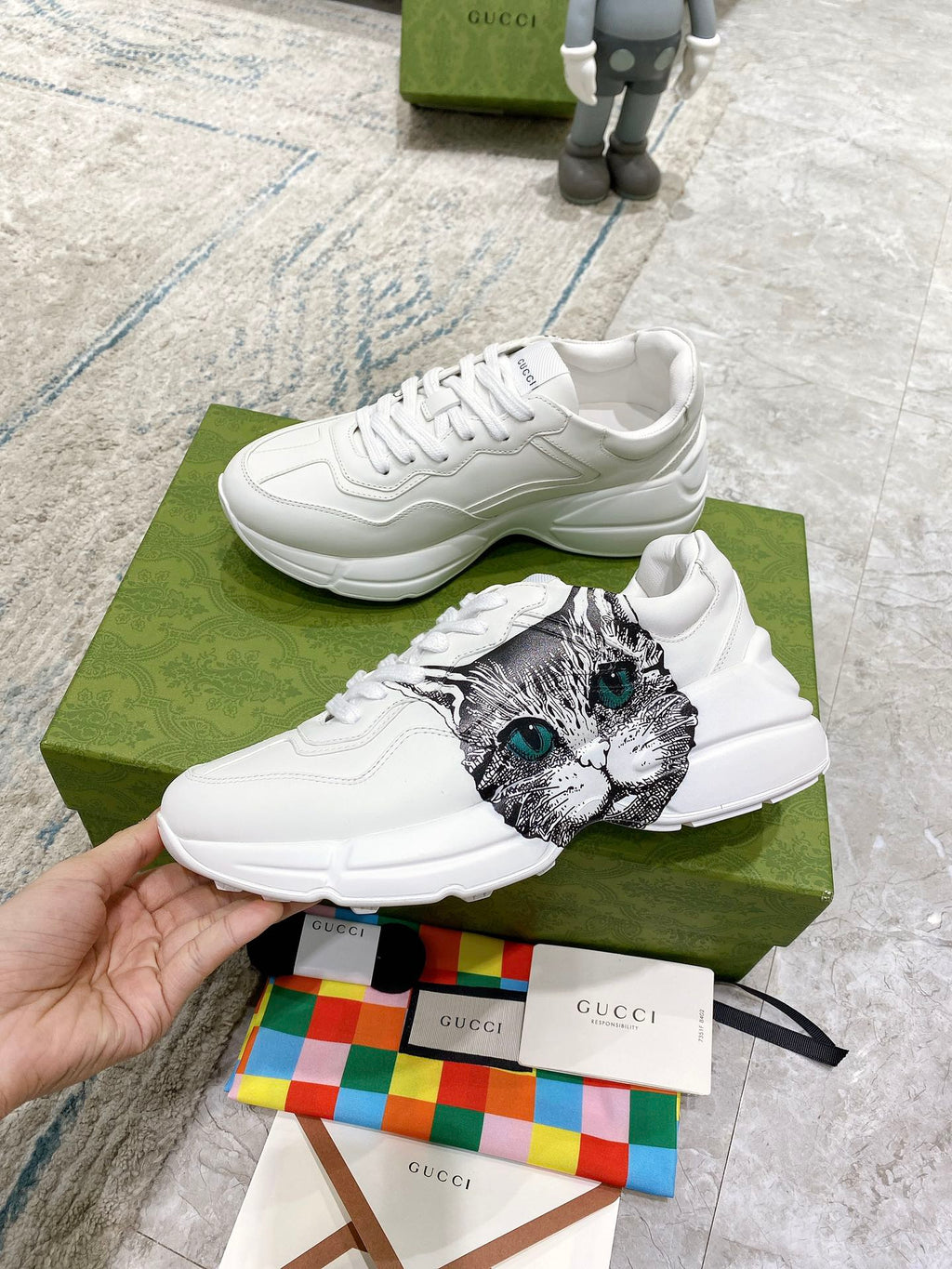 White Gucci unisex sneakers with cat-face print and green eyes; held near green box.