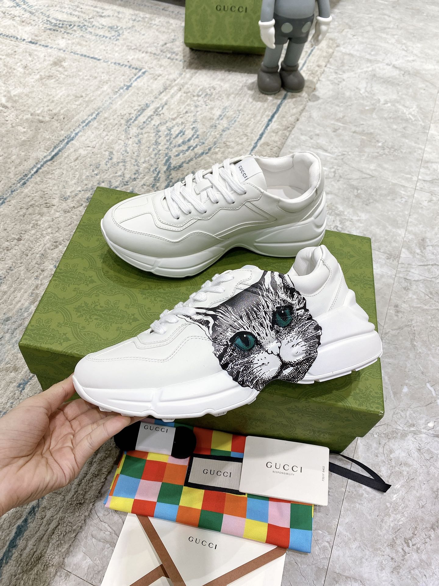 White Gucci unisex sneakers with cat-face print and green eyes; held near green box.