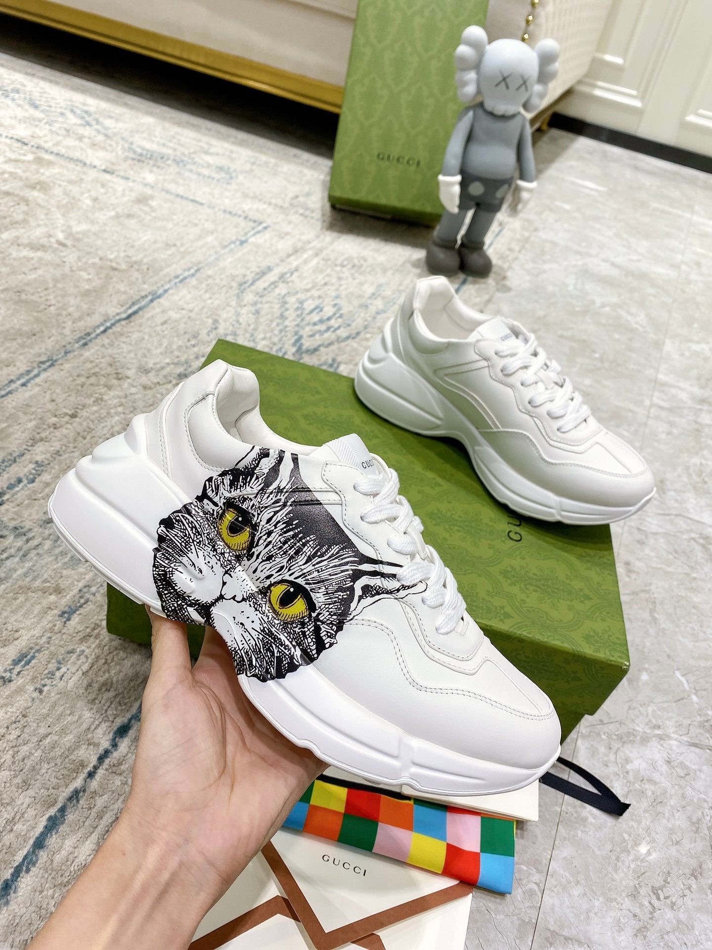 White Gucci unisex sneakers in hand; cat-face graphic on side with lace-up design.