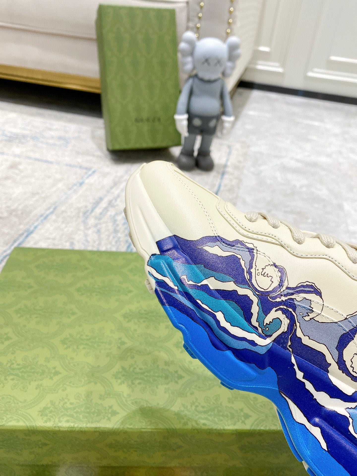 White Gucci unisex sneaker with blue wave graphic, angled on green box