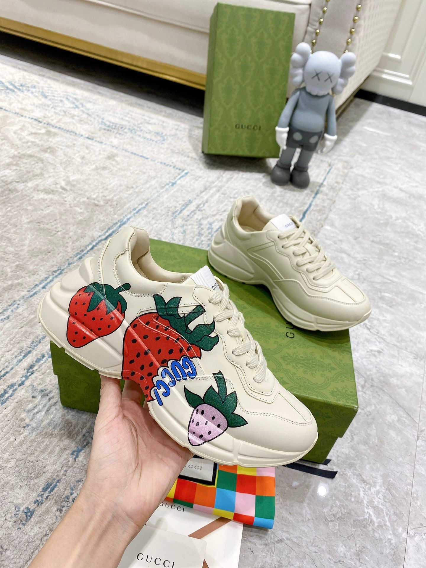 Gucci unisex sneakers held in hand: cream lace-up with strawberry and purple fruit accents.