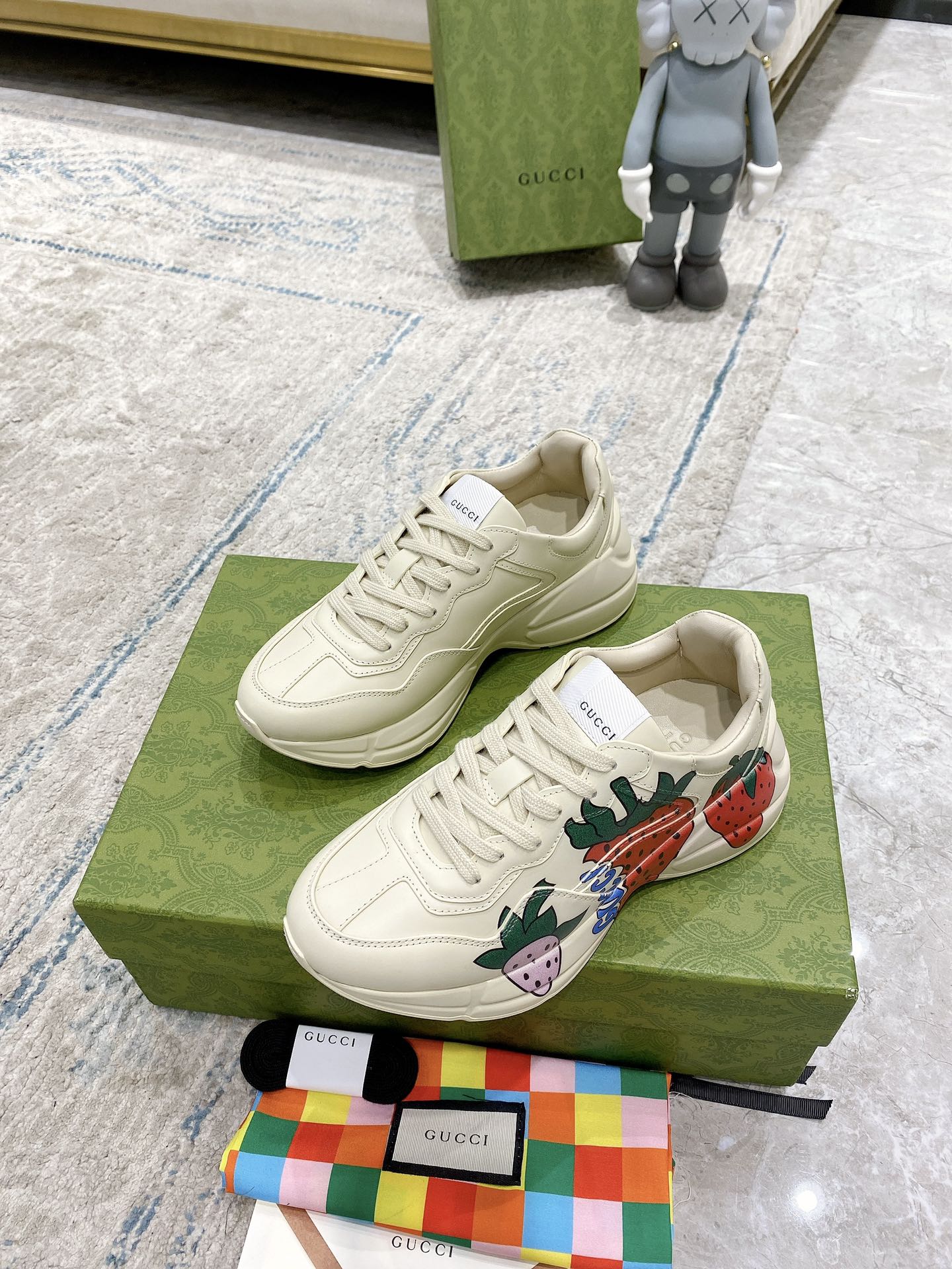 Gucci unisex sneakers on box: cream lace-up with strawberry side artwork and patterned tongue.