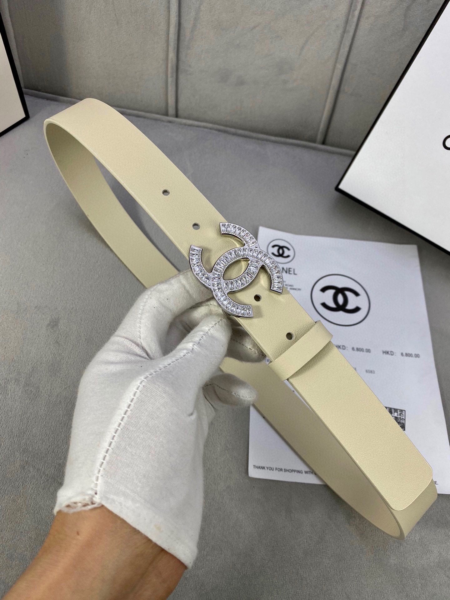 Beige leather belt with Chanel CC rhinestone buckle, held by a gloved hand