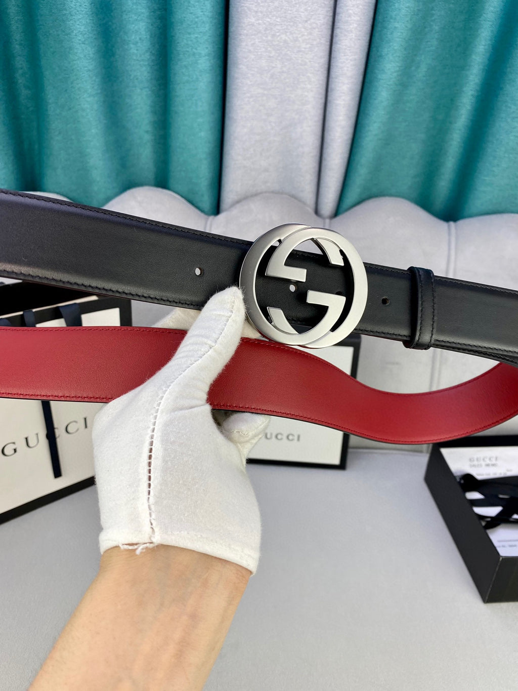 Black leather belt with silver GG buckle and red underside, held near store packaging