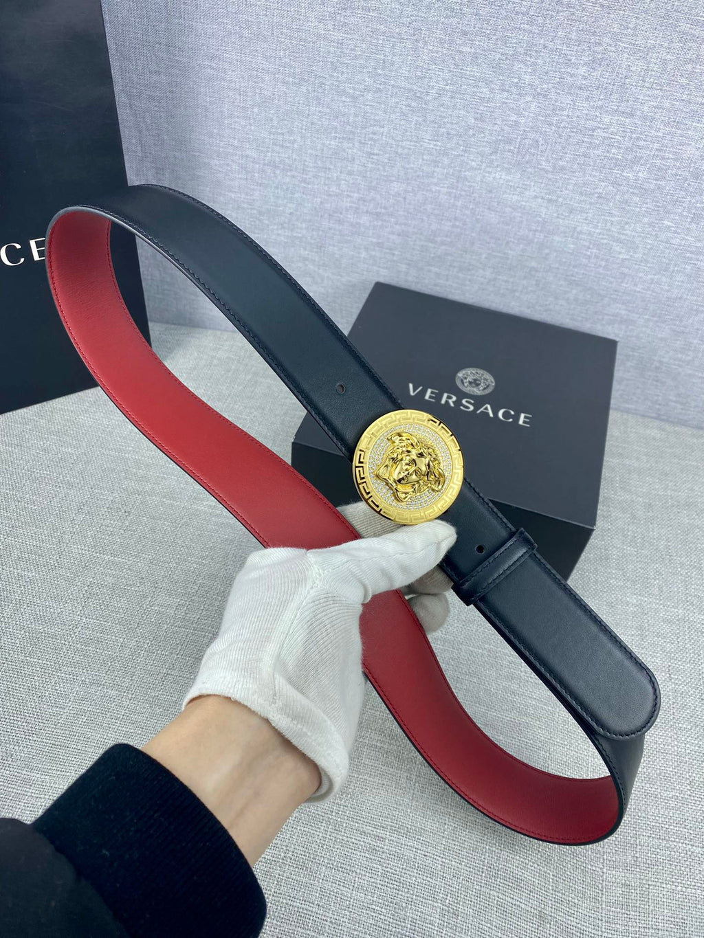 Versace leather belt held by hand: dark strap with red underside and round gold Medusa-style buckle