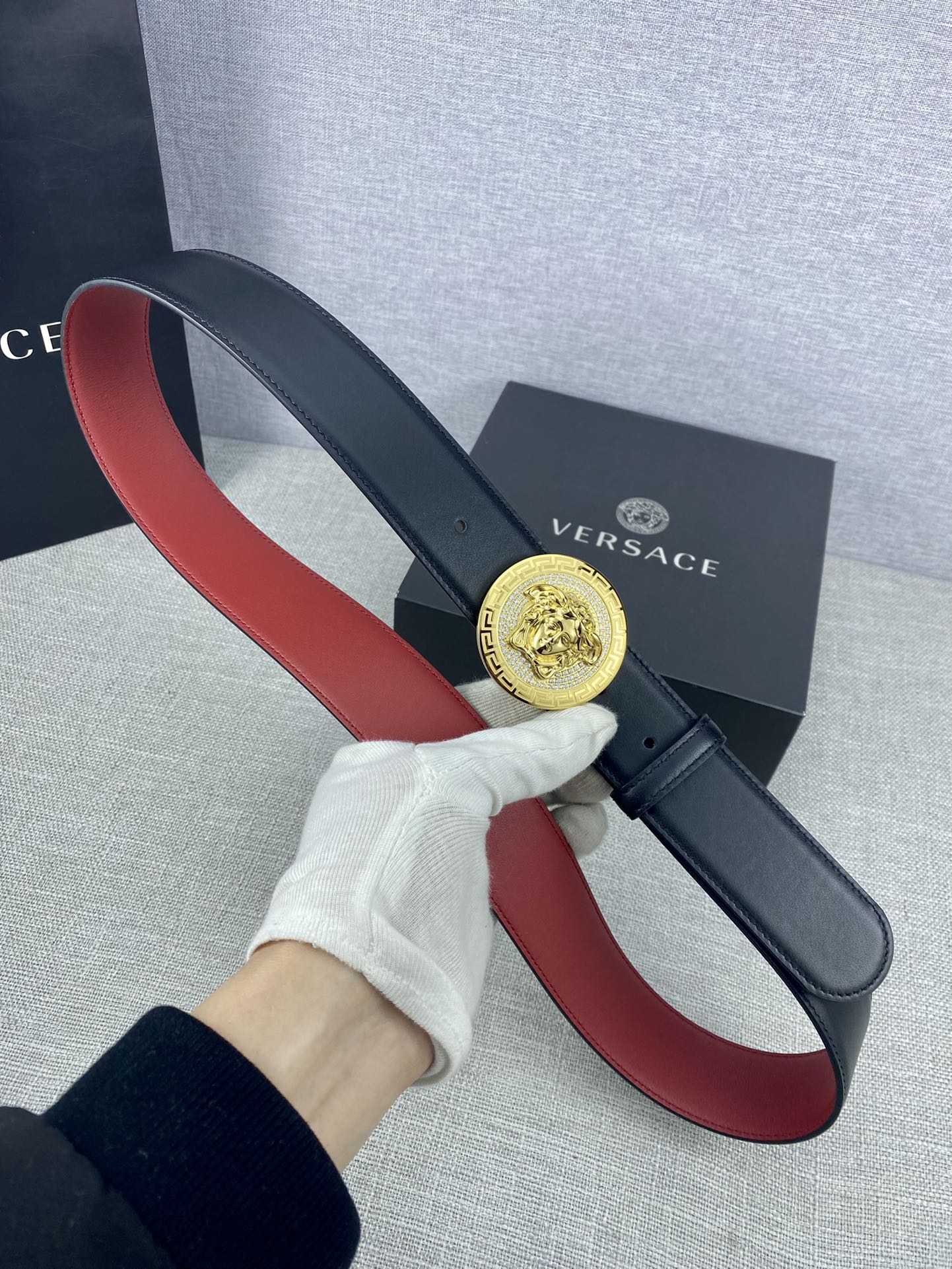 Versace leather belt held by hand: dark strap with red underside and round gold Medusa-style buckle
