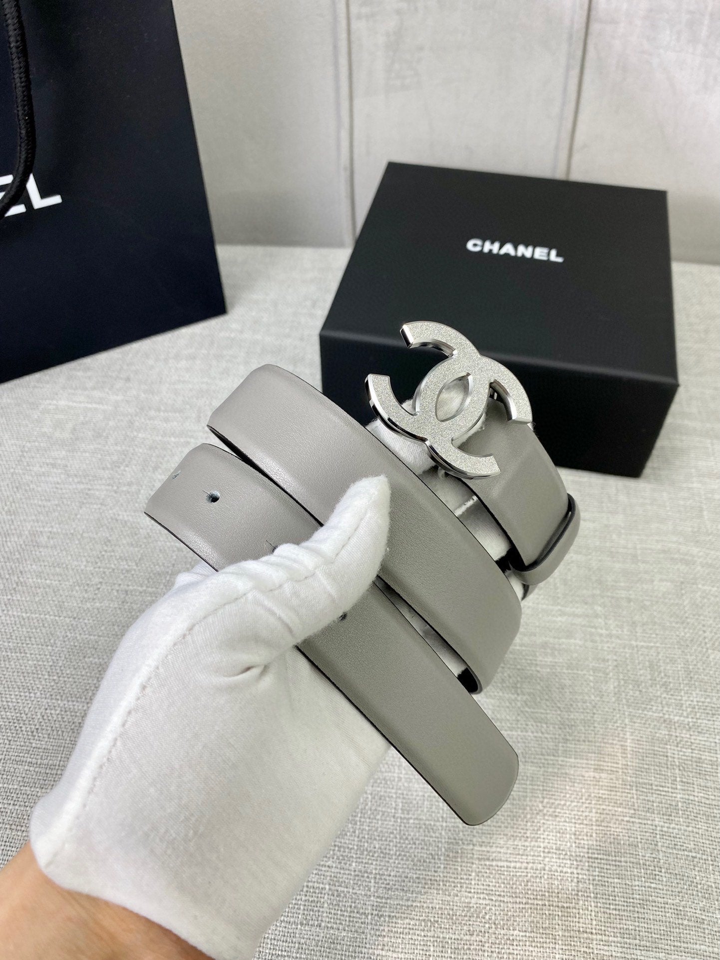 Light gray CC-buckle belt with silver logo buckle, held by white glove