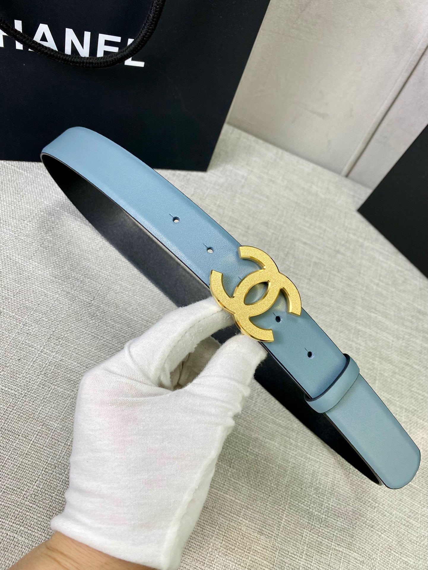Chanel belt in light blue leather with gold logo buckle, shown in gloved hand.