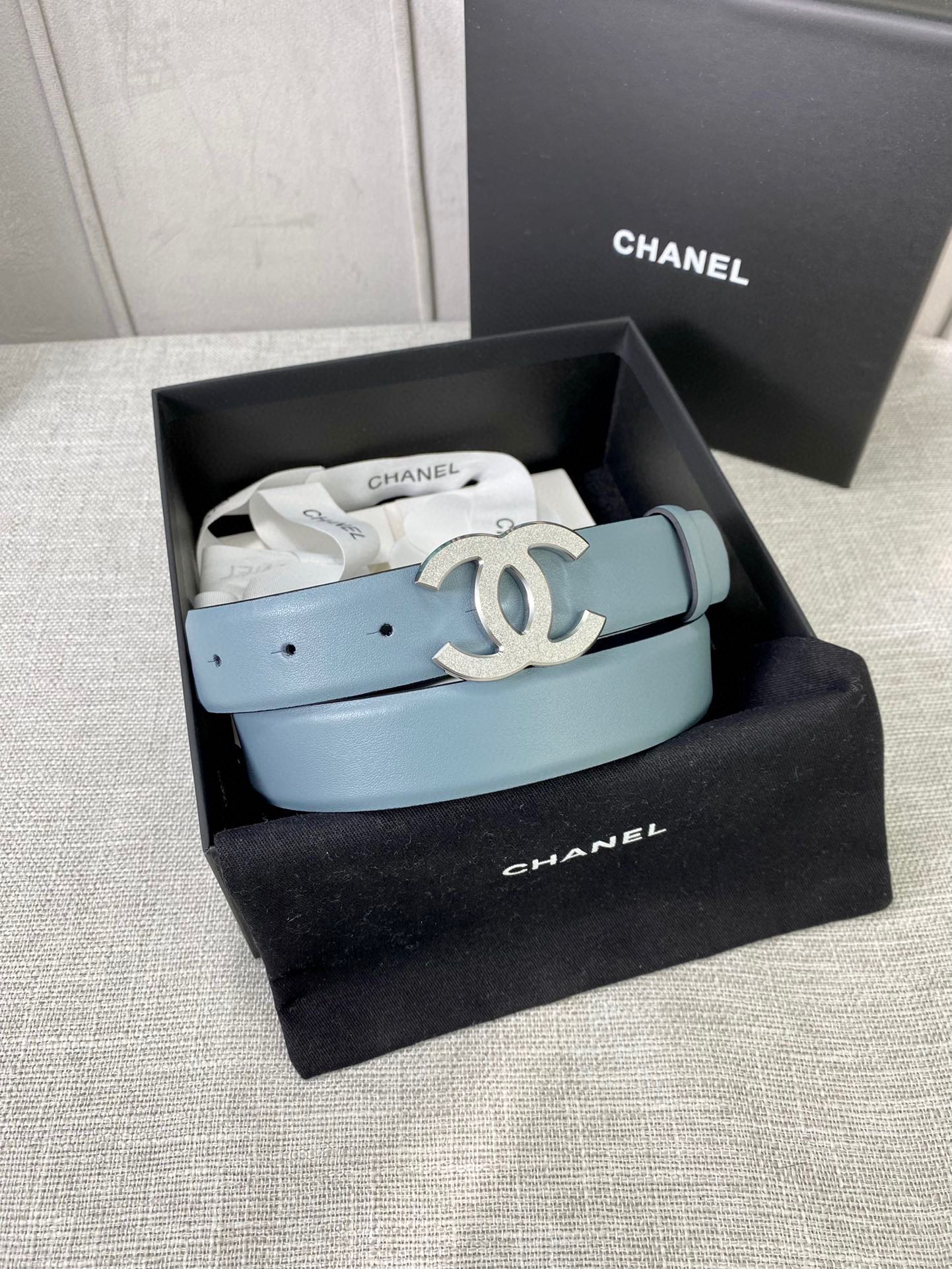 Chanel belt displayed in a gift box, light blue strap with metallic logo buckle.