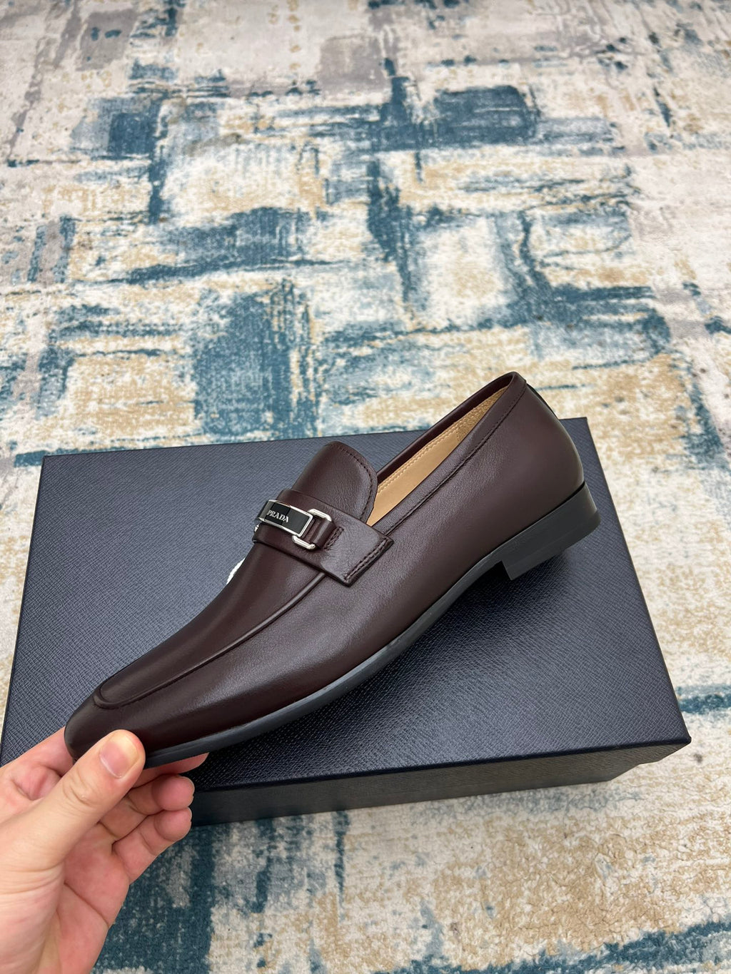 Prada unisex leather loafer with metal buckle strap, dark brown, side view on box