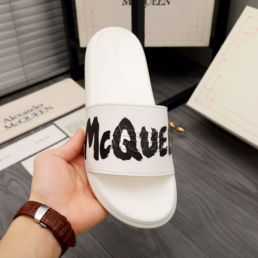 White slide sandals held in hand, black McQUEEN logo on strap