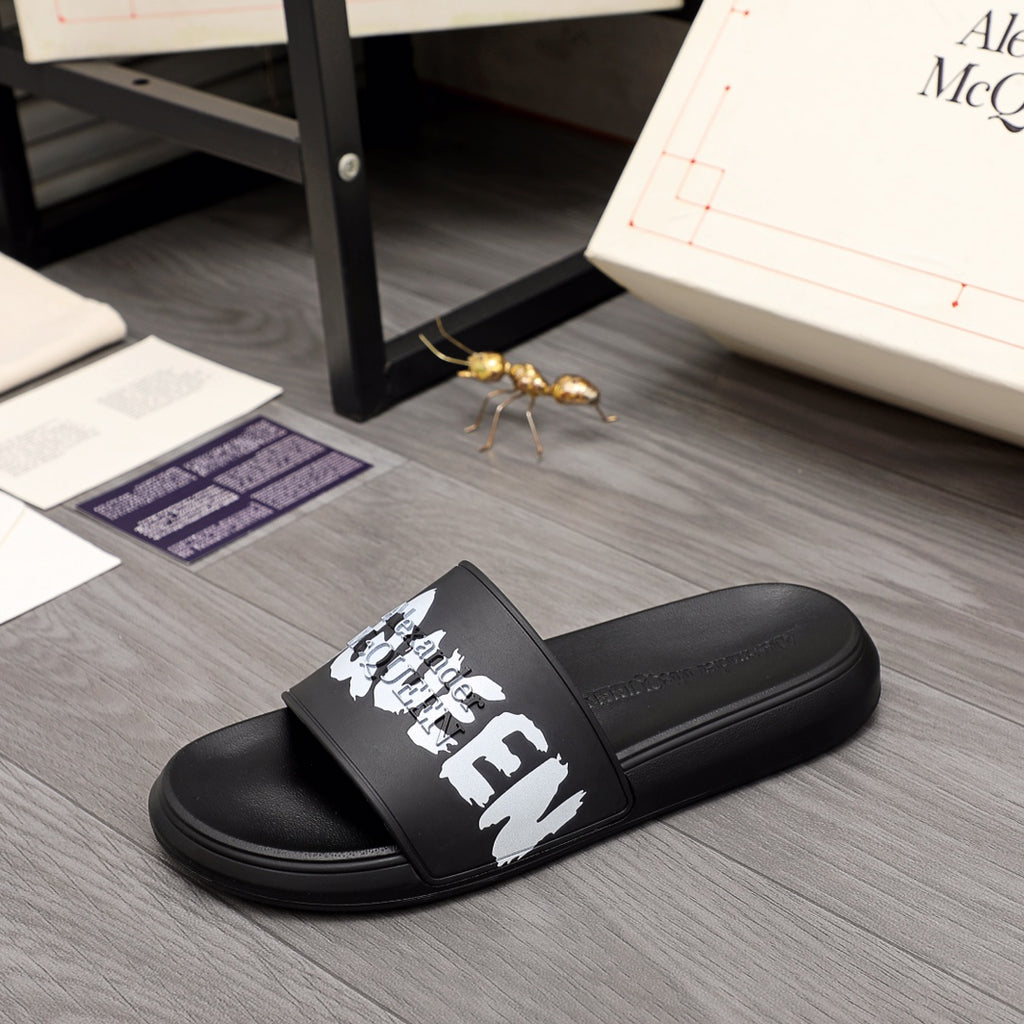 Black slide sandals with white logo graphic on the front strap, Alexander McQueen.