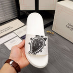 White slide sandal with black patterned emblem on the front strap, shown in hand