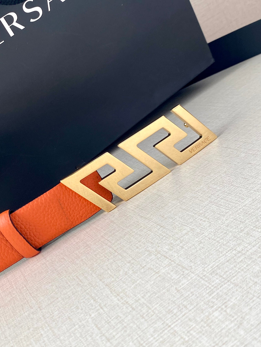 Orange leather belt with gold-tone Greca buckle, close-up detail.