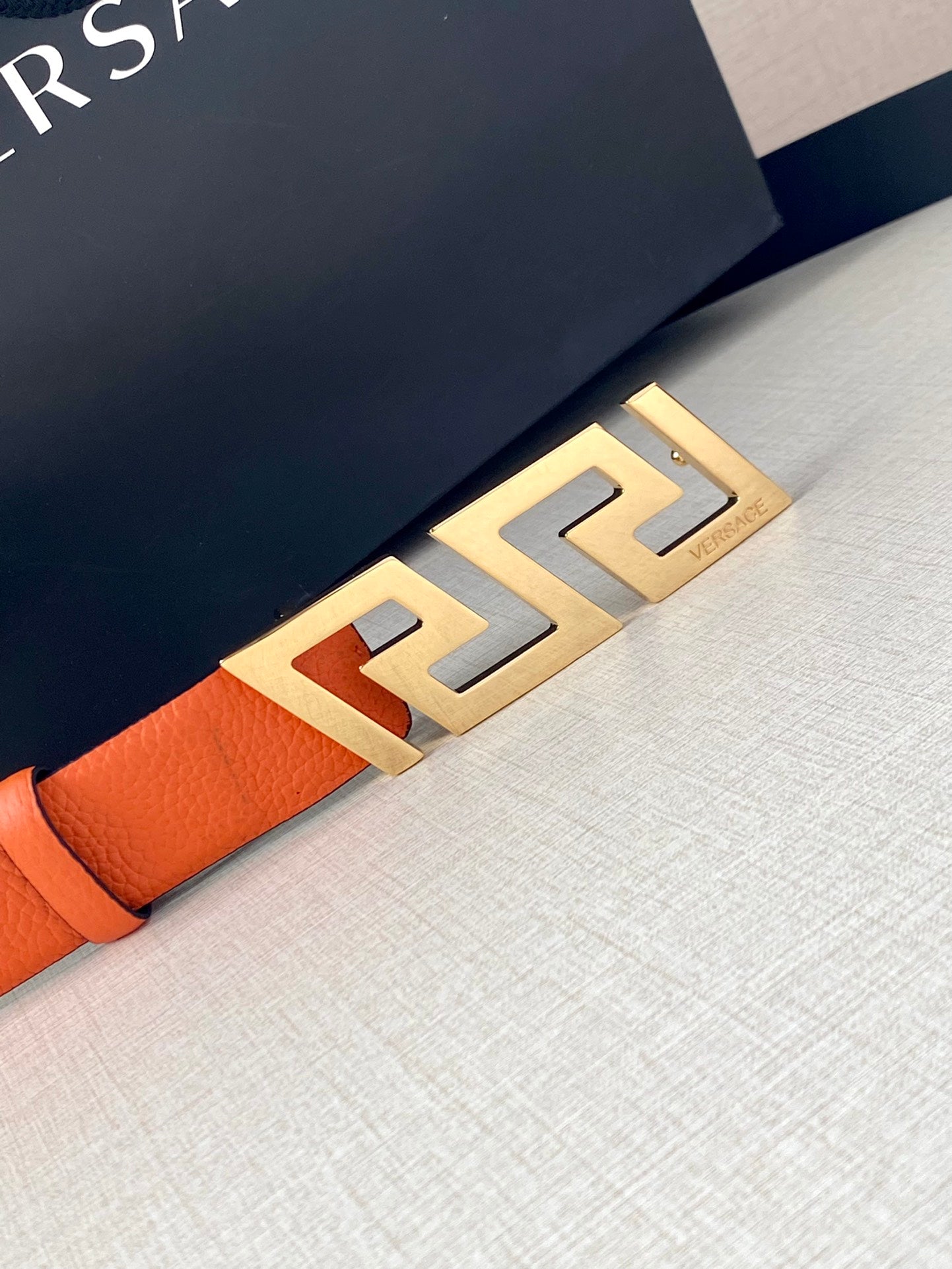 Orange leather belt with gold-tone Greca buckle, close-up detail.