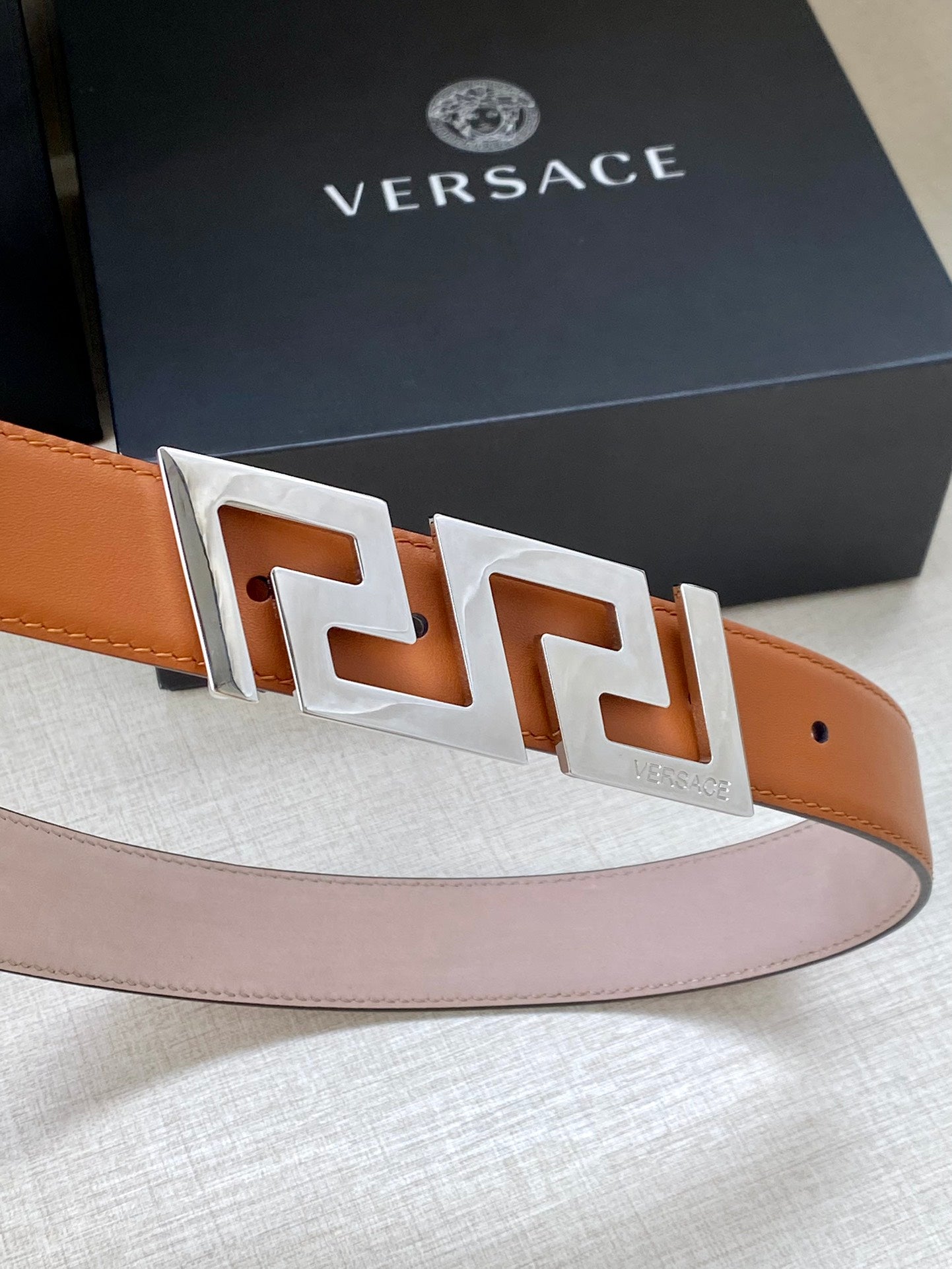 Tan leather belt with large silver-tone logo buckle and square pin holes.